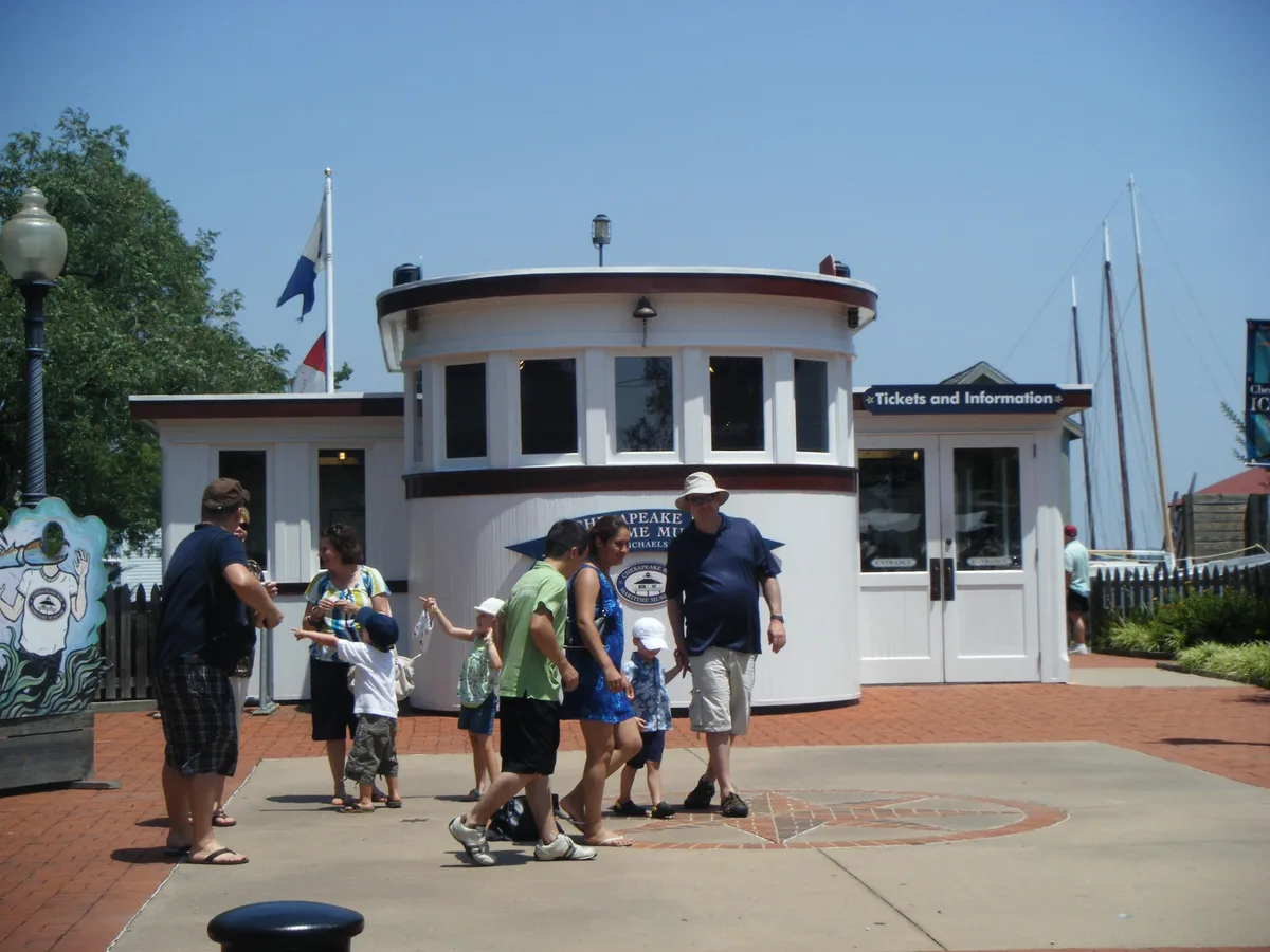 The Chesapeake Bay Maritime Museum ticket building sits in bright midday sun, with a rounded white facade, dark trim, and “Tickets and Information” sign facing a brick courtyard. Visitors cross the foreground while sailboat masts rise behind the building against a clear blue sky.