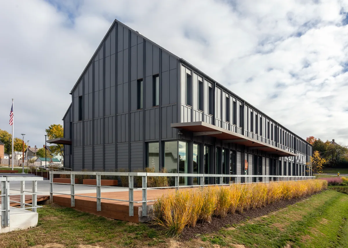 A modern dark-gray lockhouse with a steep gabled roof stands beside the canal towpath, framed by a low wooden deck, metal railings, and golden grasses in the foreground. Soft light under a cloudy sky reflects in the windows, with a flagpole and hints of historic canal buildings in the background.