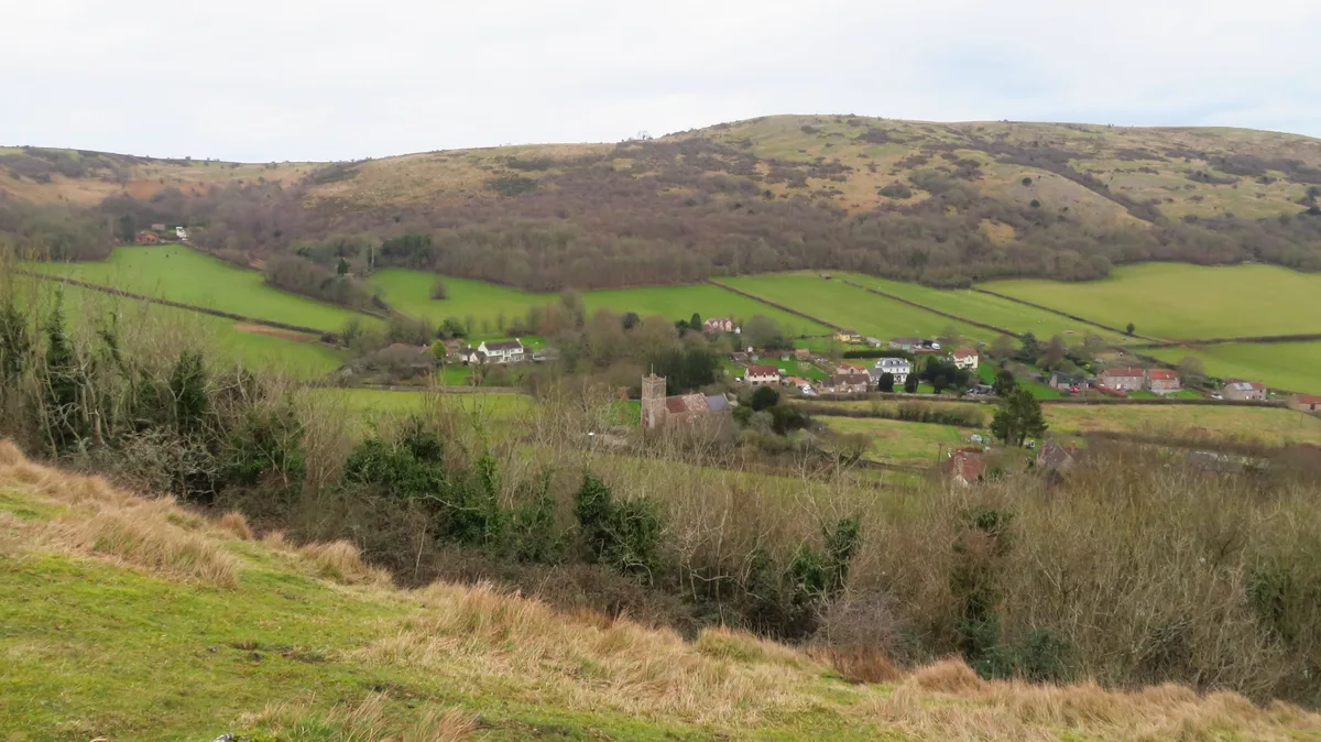 Rolling green hills and patchwork fields fill the valley below, with a small village clustered around a church tower and hedgerows dividing the landscape. Low, grassy slopes rise to a broad ridge under a pale sky, giving the overlook a muted winter look rather than golden light.