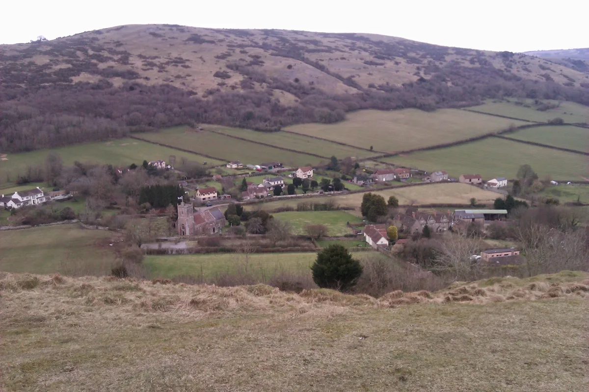 A broad view from a grassy summit ridge looks down over a patchwork of pale green fields, scattered houses, and a small stone church in the valley below. Beyond the village, a long, rounded mountain slope fills the horizon under soft, muted light.