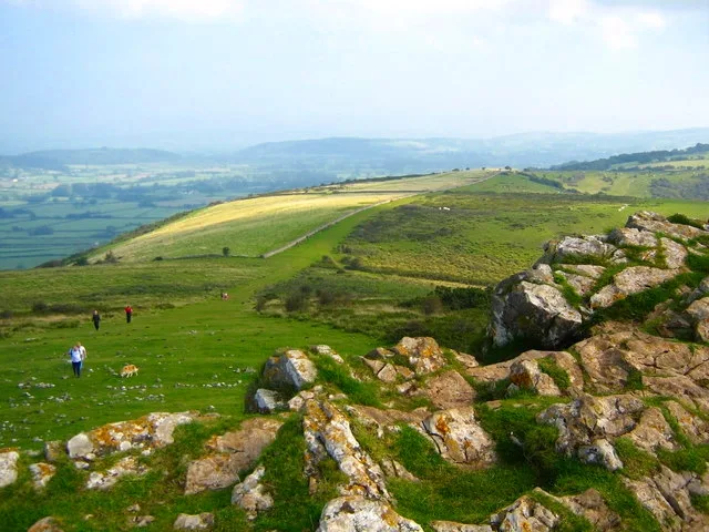 Basalt rocks fill the foreground of a grassy summit, opening to a wide view of rolling green hills and the Shenandoah Valley beyond. A few small hikers dot the slope below, while soft golden light highlights the ridge and distant haze.