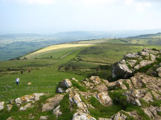 Basalt-like rock outcrops fill the foreground, with a grassy ridge dropping away to a broad Shenandoah Valley panorama under soft golden light. A few small hikers dot the slope, and hazy blue hills layer the horizon beyond Front Royal.