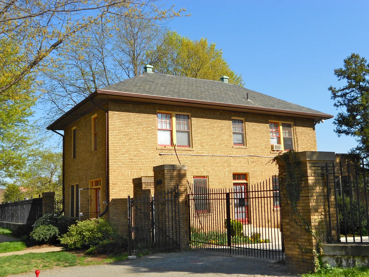 A two-story yellow-brick building with a dark hipped roof sits behind a black iron gate and brick pillars, framed by spring-green trees under a clear blue sky. The scene feels quiet and historic, with sunlight catching the warm brick and red-trimmed windows.