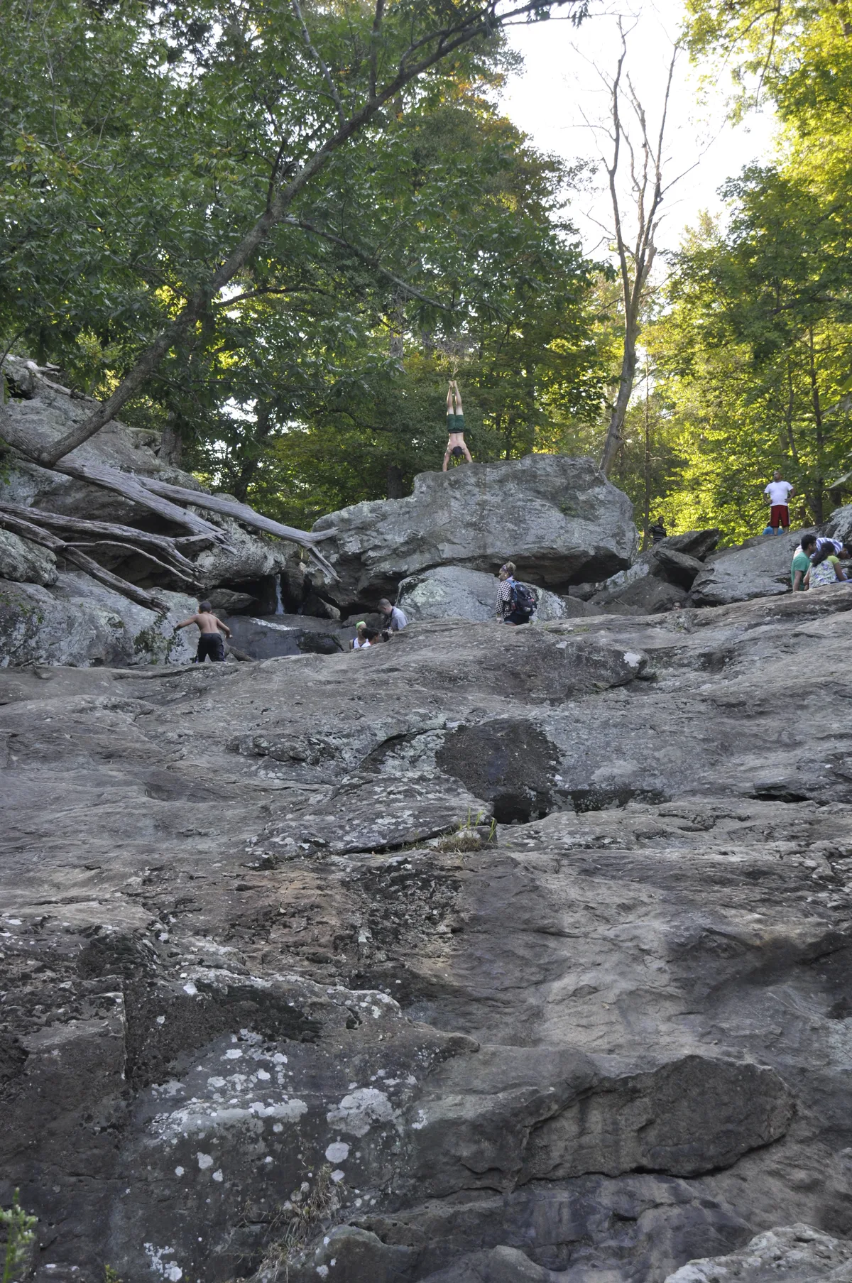 Steep gray granite slabs rise through a shaded forest at Cunningham Falls, with a person balancing in a handstand on a large boulder near the top. Several visitors stand and climb among the rocks under leafy trees lit by soft late-afternoon sun.