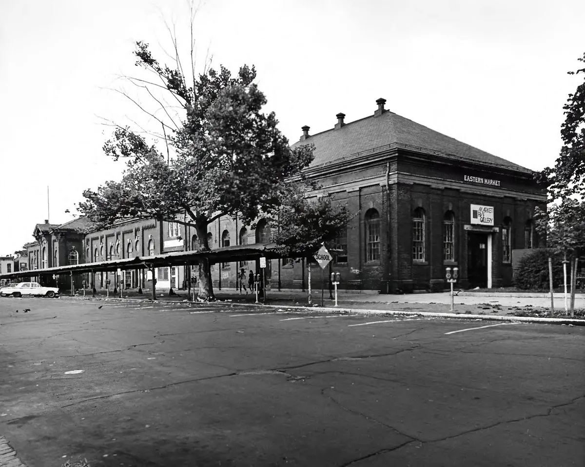Black-and-white view of Eastern Market’s brick facade and hipped roof, with “Eastern Market” and “Market & Gallery” signs over the entrance. A long covered walkway lines the side of the building, framed by trees and an empty cracked parking lot in the foreground.