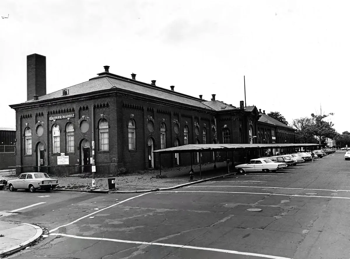 Historic Eastern Market’s long brick facade fills the frame, with arched windows, a steep hipped roof, and a tall chimney rising above the Capitol Hill street. Vintage cars line the curb under a low awning, and the wide intersection in front is marked by bright painted road lines.