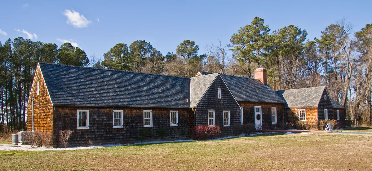 A long, low cedar-shingled lodge with a steep gray roof sits in a sunny clearing, framed by tall pines and bare winter trees. Warm golden light highlights the rustic siding and brick chimney under a clear blue sky.