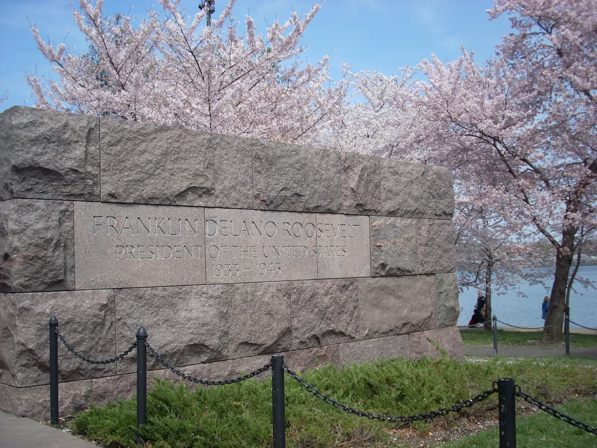 A rough-hewn granite wall engraved “Franklin Delano Roosevelt, President of the United States, 1933–1945” stands in the foreground, with pale pink cherry blossoms filling the background. The scene opens toward the water at right, with spring foliage and black chain posts framing the memorial under a clear blue sky.