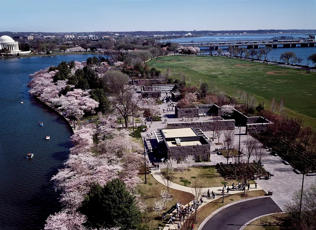High aerial view of the Franklin Delano Roosevelt Memorial along the Tidal Basin, with spring cherry blossoms lining the curved shoreline beside the granite terraces and walkways. The Jefferson Memorial, bridges, and calm blue water frame the memorial’s layered stone rooms and open green lawn.
