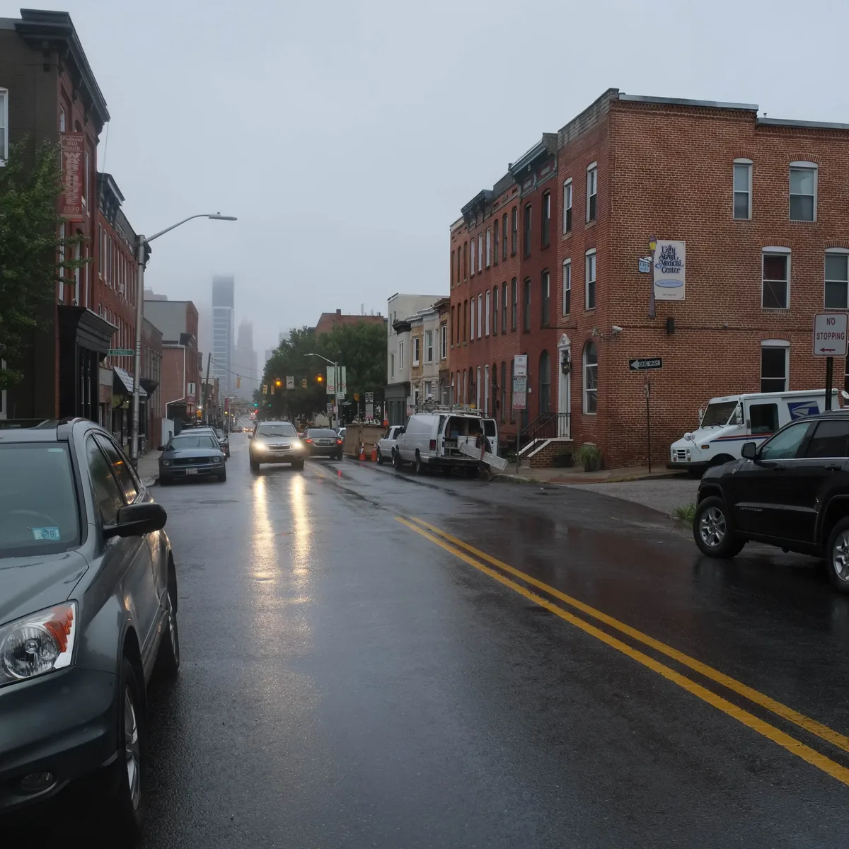 Wet Federal Hill street lined with brick rowhouses and parked cars, looking toward the hazy Baltimore skyline in the distance. Reflections from car headlights streak across the rain-dark pavement under a dim blue-hour sky.
