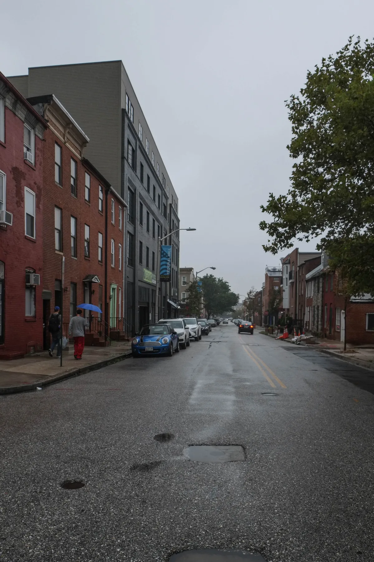 A wet, gray street in Federal Hill stretches into the distance beneath an overcast sky, lined with red brick rowhouses, parked cars, and a few trees. Two pedestrians walk along the left sidewalk, and the damp pavement reflects the muted blue-hour light.