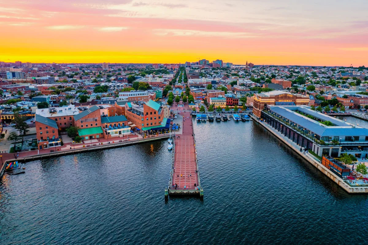 Golden hour lights Fell’s Point’s brick rowhouses and waterfront buildings beside a long pier stretching into the harbor. The calm water reflects the pink and gold sky above the dense city grid.