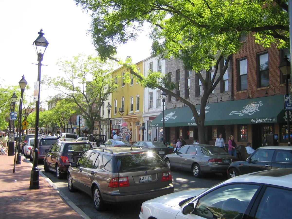 Sunlit Fells Point street lined with leafy trees, black gas-style lampposts, and parked cars in front of colorful brick rowhouses and storefronts. A green awning for The Greene Turtle anchors the block, giving the cobblestone waterfront district a lively neighborhood feel.