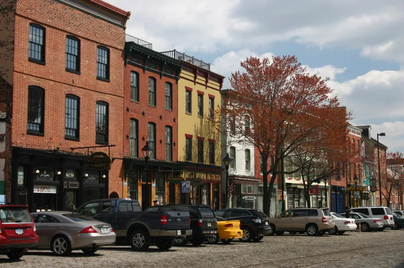 Historic brick rowhouses and storefronts line a cobblestone street in Fells Point, with parked cars along the curb and a large autumn tree softening the block. The scene has a warm late-day feel under a partly cloudy sky, emphasizing the neighborhood’s old waterfront character.