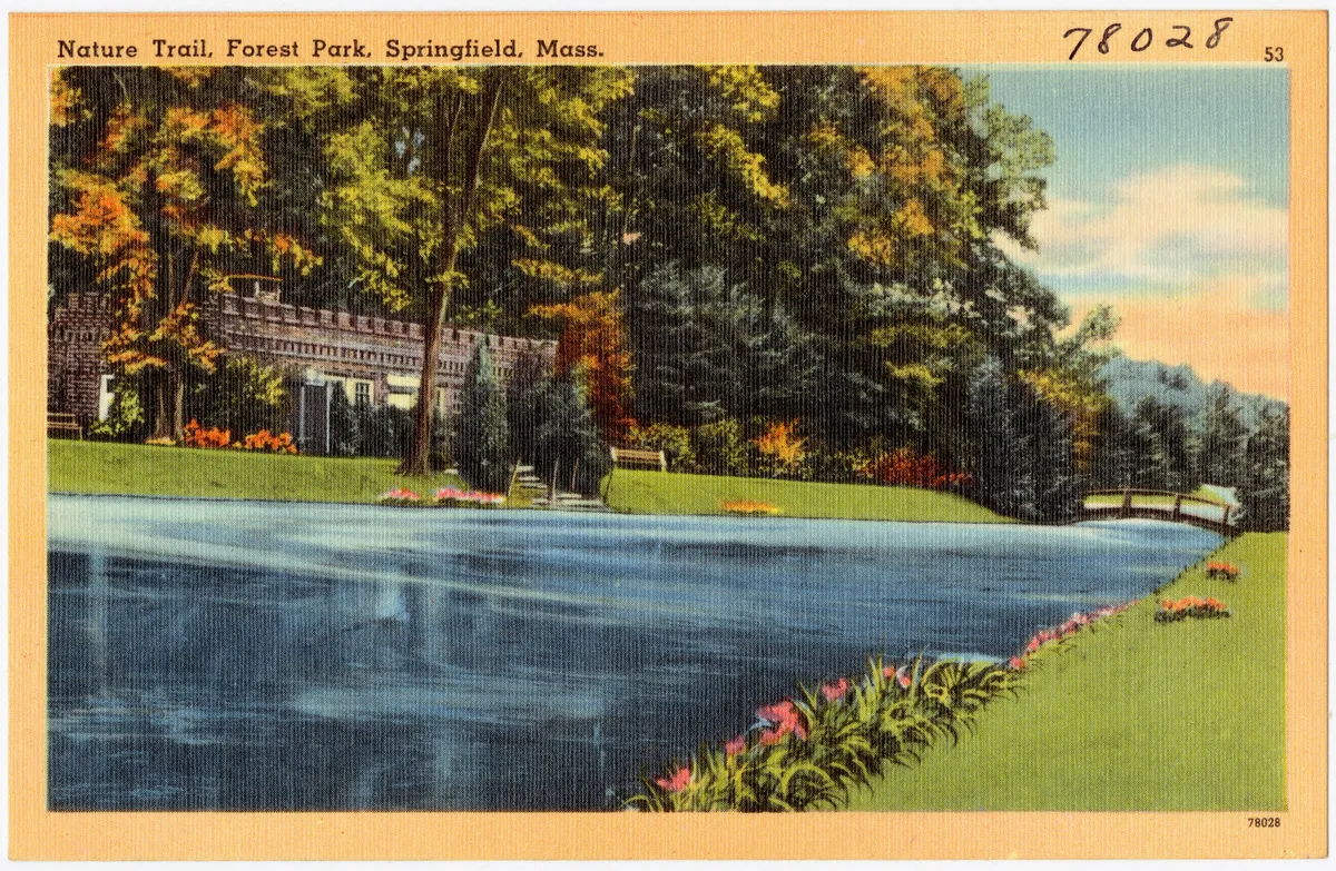 A calm, dark-blue waterway curves through a manicured park, bordered by bright green lawns, flower beds, and dense trees in warm late-day light. A low stone-and-brick building sits back on the left, with a small bridge visible in the distance under a soft pastel sky.