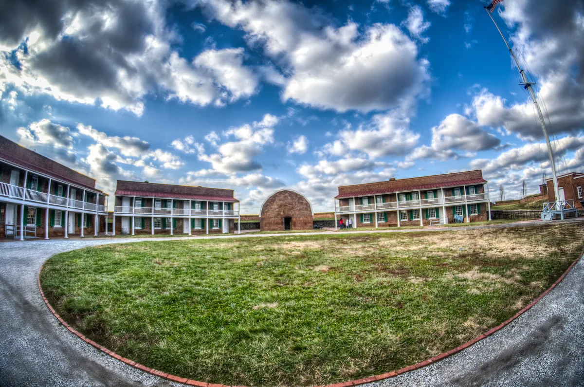 Wide view inside Fort McHenry’s star-shaped courtyard, with low brick barracks and white balconies curving around a grassy parade ground. A dramatic sky with bright, scattered clouds fills the frame, and a tall flagpole rises at the right edge.