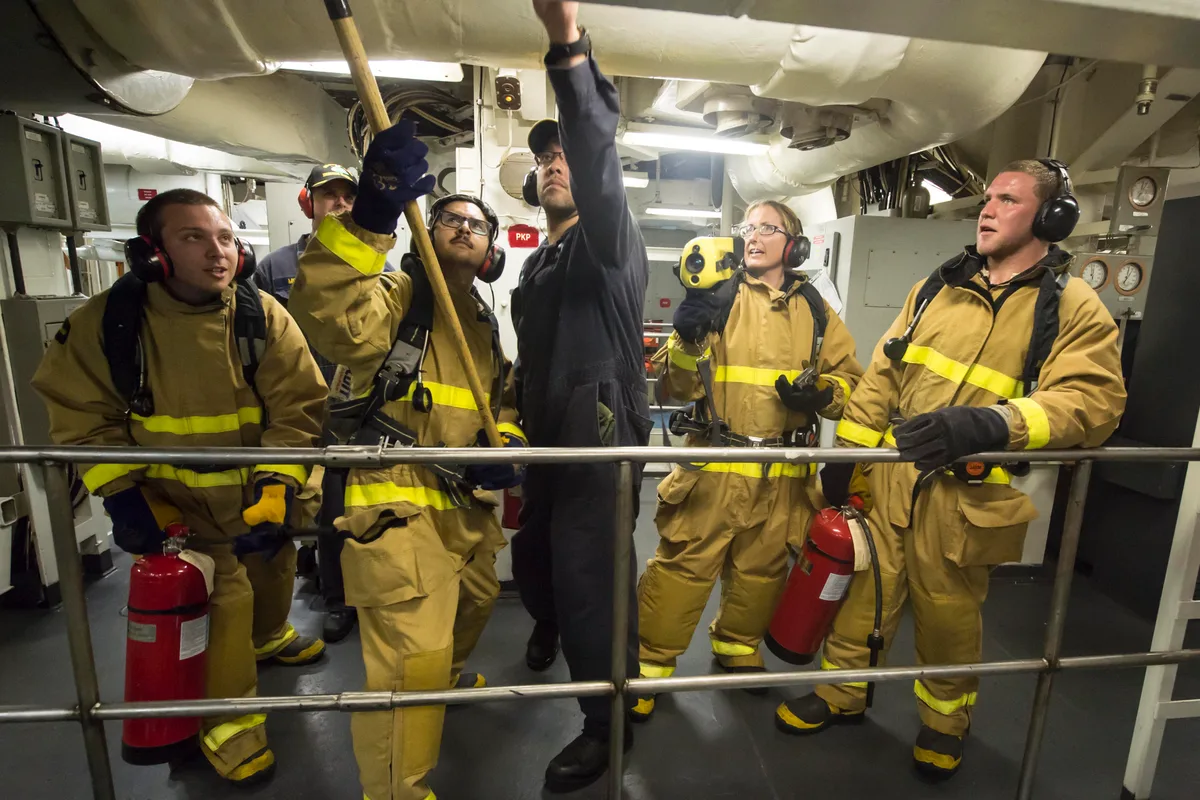 Several crew members in tan firefighting gear and headsets stand shoulder to shoulder on a ship’s deck, with one reaching up to a ceiling pipe and others holding flashlights and fire extinguishers. Bright overhead lighting and tight railings give the scene a cramped, industrial look.