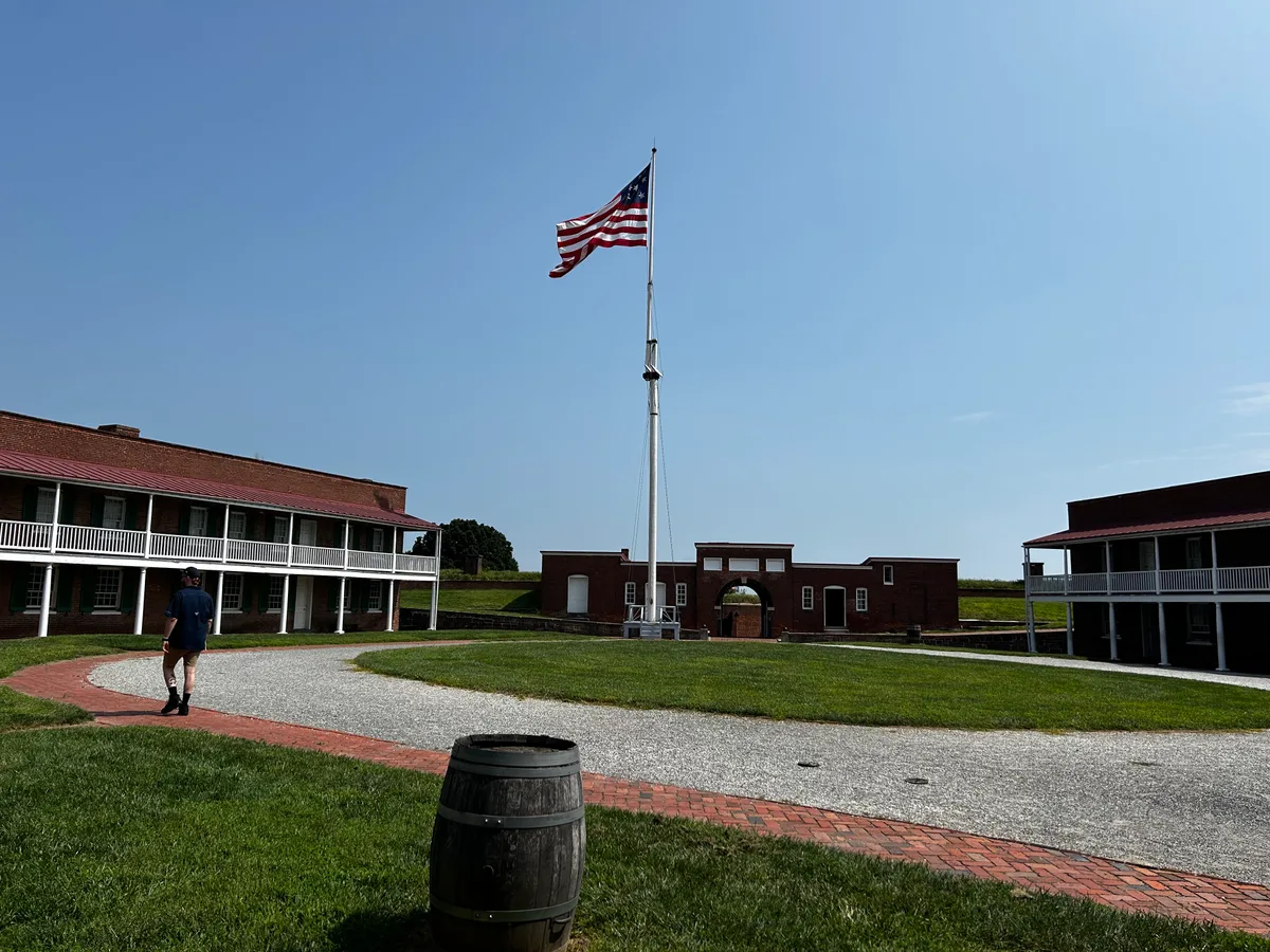 Inside Fort McHenry’s grassy courtyard, a tall flagpole with a waving American flag rises above a circular gravel path, brick barracks, and a central arched gateway. Bright clear light and a wide blue sky emphasize the fort’s open, historic interior layout.