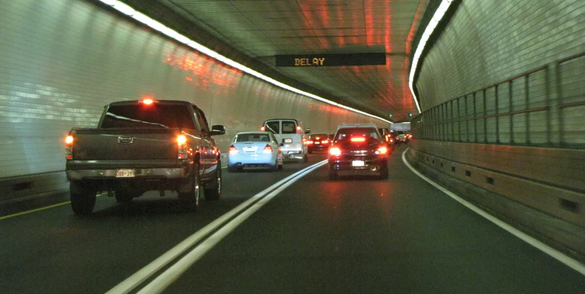 Traffic moves through a tiled tunnel beneath Fort McHenry, with cars and pickup trucks following the curved lanes under a glowing “DELAY” sign. The greenish walls and overhead lighting create a long, enclosed perspective with red brake lights reflecting on the roadway.
