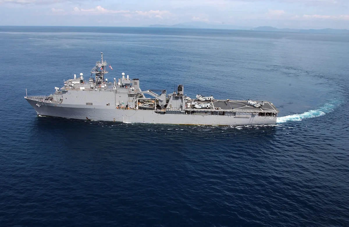 An amphibious dock landing ship cruises through calm open water, its gray hull and helicopter deck cutting a bright white wake behind it. The wide aerial view shows the vessel in profile against a deep blue sea and hazy horizon.