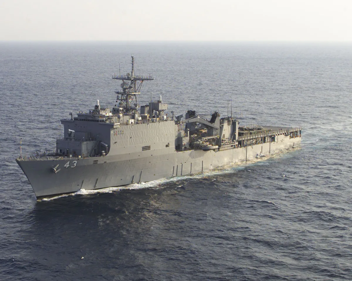 A gray naval ship with hull number 43 cuts through calm open water, its deck cranes and radar mast rising above the flat, sunlit sea. The vessel is shown in a wide side view, with a bright horizon and faint wake trailing along its hull.
