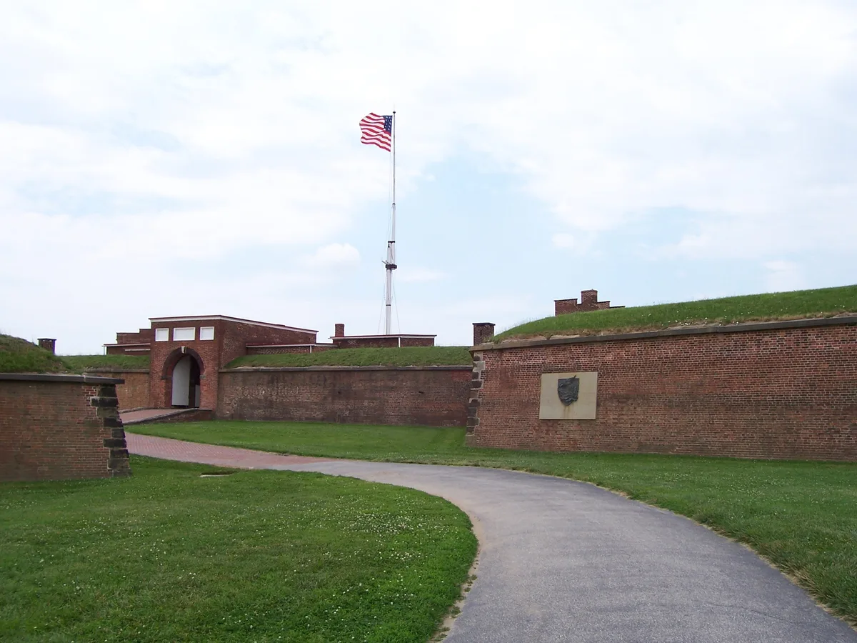A curved paved path leads through Fort McHenry’s grassy brick ramparts to a low arched entrance, with the American flag flying on a tall mast above the fort walls. Soft daylight and a pale sky highlight the historic masonry and earthworks.