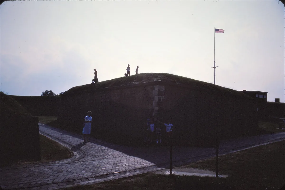 Fort McHenry’s grassy, brick-lined star-shaped ramparts are silhouetted against a bright evening sky, with a tall American flag flying from the fort’s peak. A few visitors stand along the slope and walkway, adding scale to the low, sweeping profile of the historic fort.