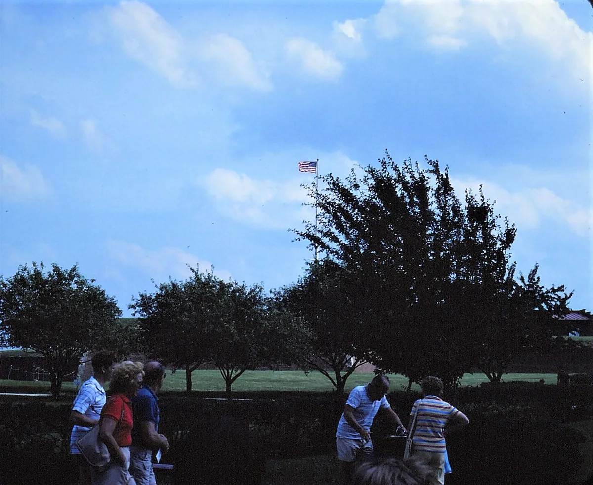 Visitors stand in the foreground beneath dark tree silhouettes, looking toward Fort McHenry’s grassy grounds and a U.S. flag flying high against a bright blue sky with scattered clouds. The scene feels late-day and quiet, with the fort’s open lawn and harbor-side setting stretching into the distance.
