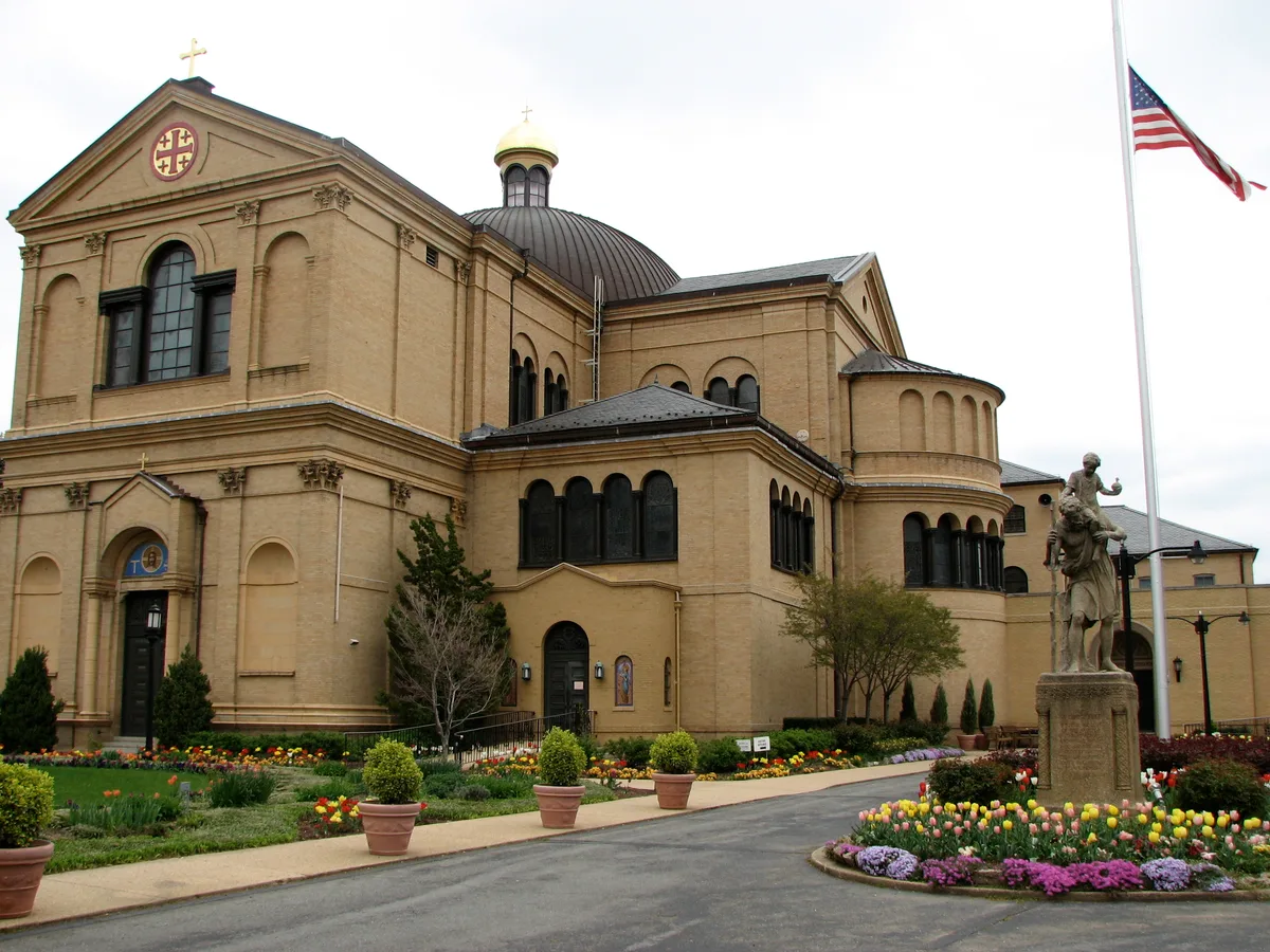 The Franciscan Monastery’s tan stone basilica rises above a landscaped garden with spring tulips, potted shrubs, and a statue near the flagpole. Its rounded domes, arched windows, and cross-topped roofline stand out against the overcast midday sky.