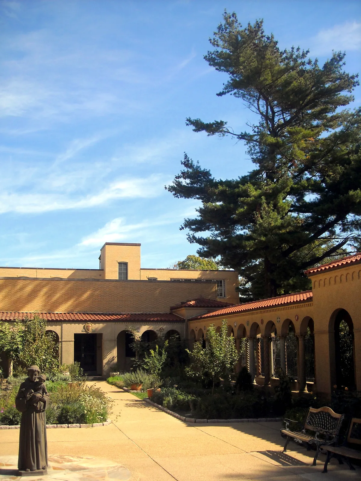 Sunlit monastery courtyard with a tan stucco cloister, red-tile roof, and arched colonnade framing a small garden path. A statue stands in the foreground beside low plantings, with a tall pine and bright blue sky filling the upper half of the scene.