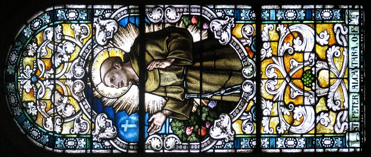 A stained-glass panel shows St. Peter in a brown robe, haloed and framed by elaborate white scrollwork, with blue and gold jewel tones glowing around him. The narrow window is set in a dark interior and includes the vertical inscription “ST. PETER FALC. TRAF.” along the right edge.