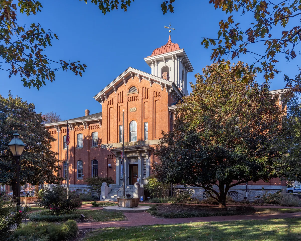 Frederick City Hall’s red-brick Victorian façade rises behind a small landscaped plaza, with arched windows, ornate trim, and a tower topped by a copper-colored roof and weather vane. Golden-hour light warms the building while tree branches frame the clear blue sky above.