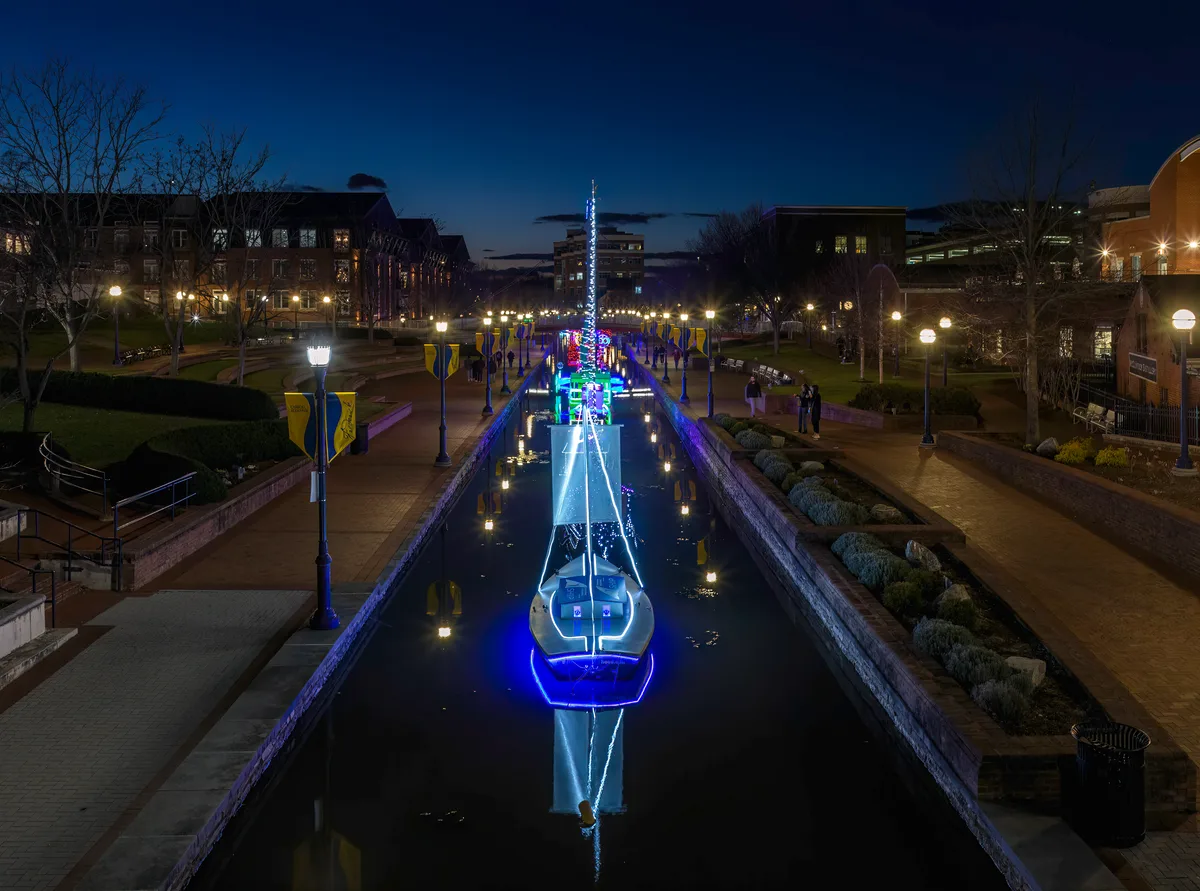 A brightly lit boat display floats in the narrow Carroll Creek canal at dusk, its blue and white lights reflecting in the water beneath a darkening sky. Brick walkways, lampposts, and modern historic-district buildings line both sides of the canal, with a few people strolling nearby.