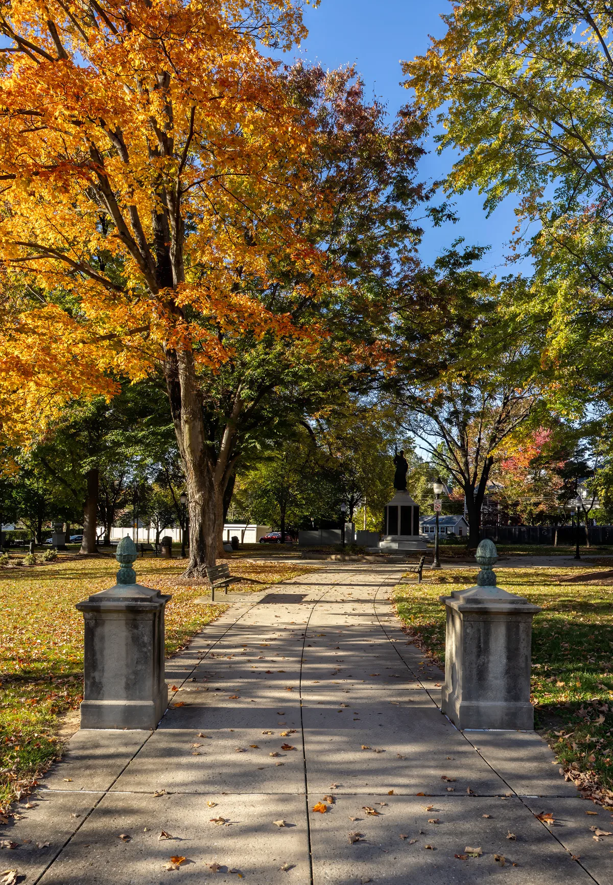 A tree-lined park path leads toward a dark statue on a pedestal, framed by ornamental stone posts and benches under vivid autumn foliage. Golden sunlight warms the orange, yellow, and green leaves against a clear blue sky, with fallen leaves scattered across the sidewalk.