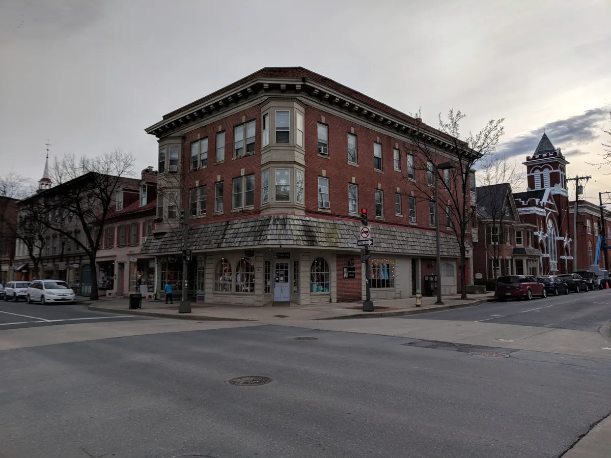 A wide street-corner view of downtown Frederick shows a three-story red-brick corner building with bay windows and a shingled storefront base, framed by older shopfronts and leafless trees. Across the intersection, a red-and-white church-like building with a pointed tower adds to the historic streetscape under a pale evening sky.