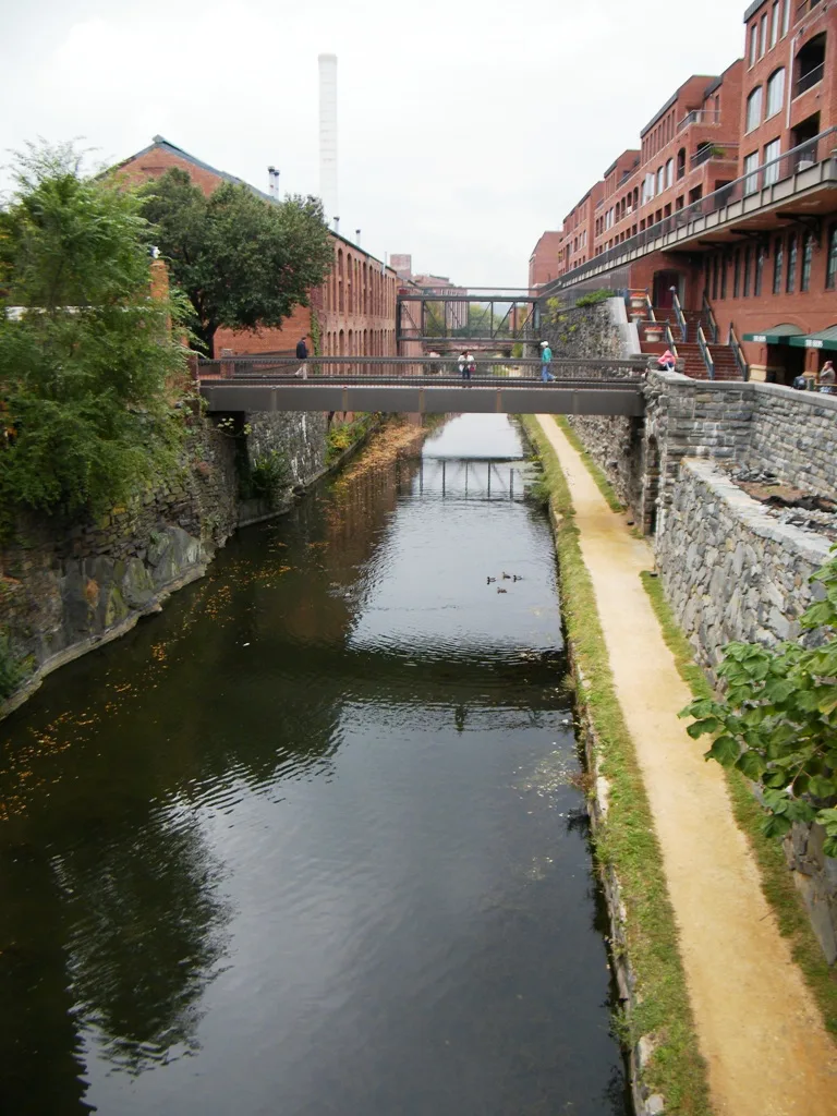 The C&O Canal runs between brick Georgetown buildings and stone embankments, with a pedestrian bridge crossing the water and its dark surface reflecting the overcast sky. A narrow towpath follows the right bank, where a few people walk beside the historic canal.