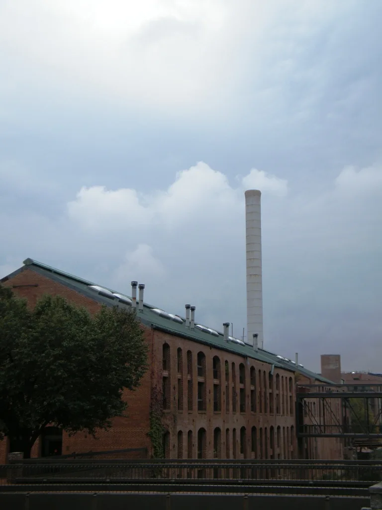 Brick industrial buildings line the C&O Canal towpath, with a tall white smokestack rising behind arched windows and a leafy tree in the foreground. Dark railings and a footbridge frame the lower edge under a hazy, bright sky.