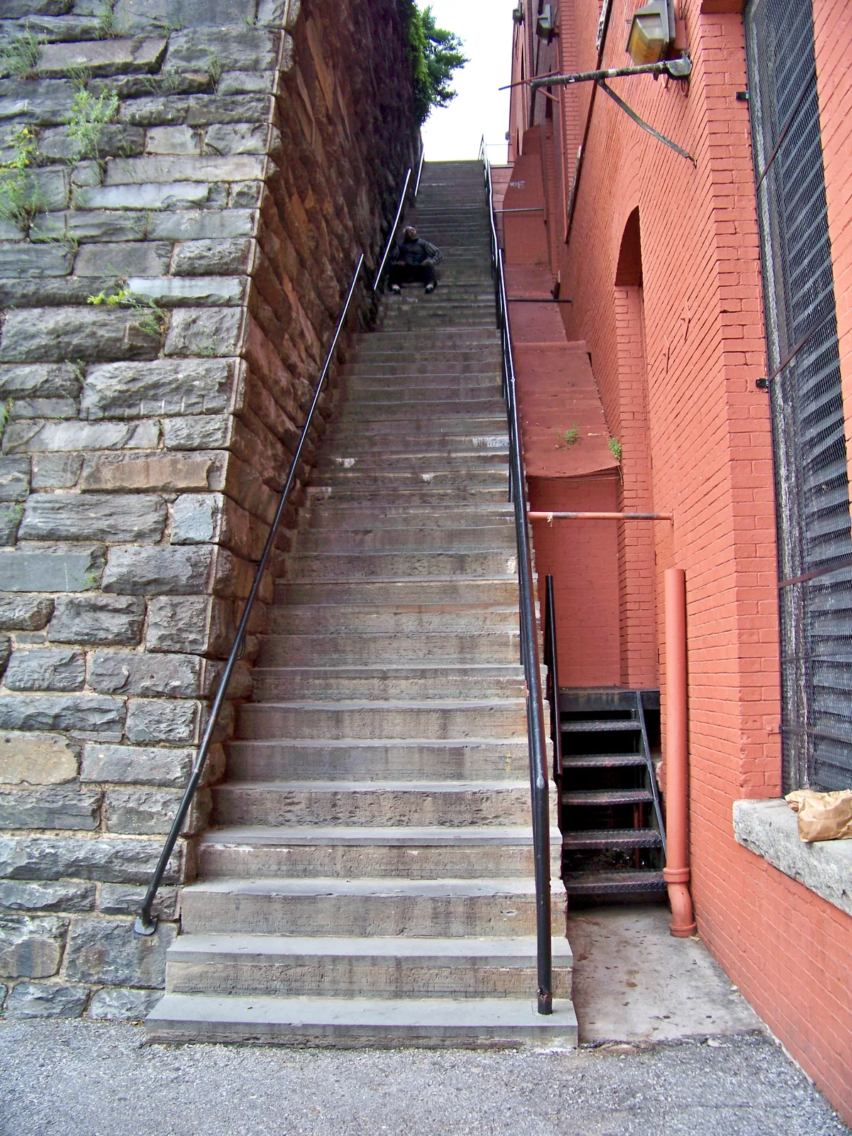Steep concrete stairs rise between a rough gray stone retaining wall and a red brick building with black railings, creating a tight, vertical urban frame. A small figure sits partway up the steps, emphasizing the height and narrowness of the Exorcist Steps.