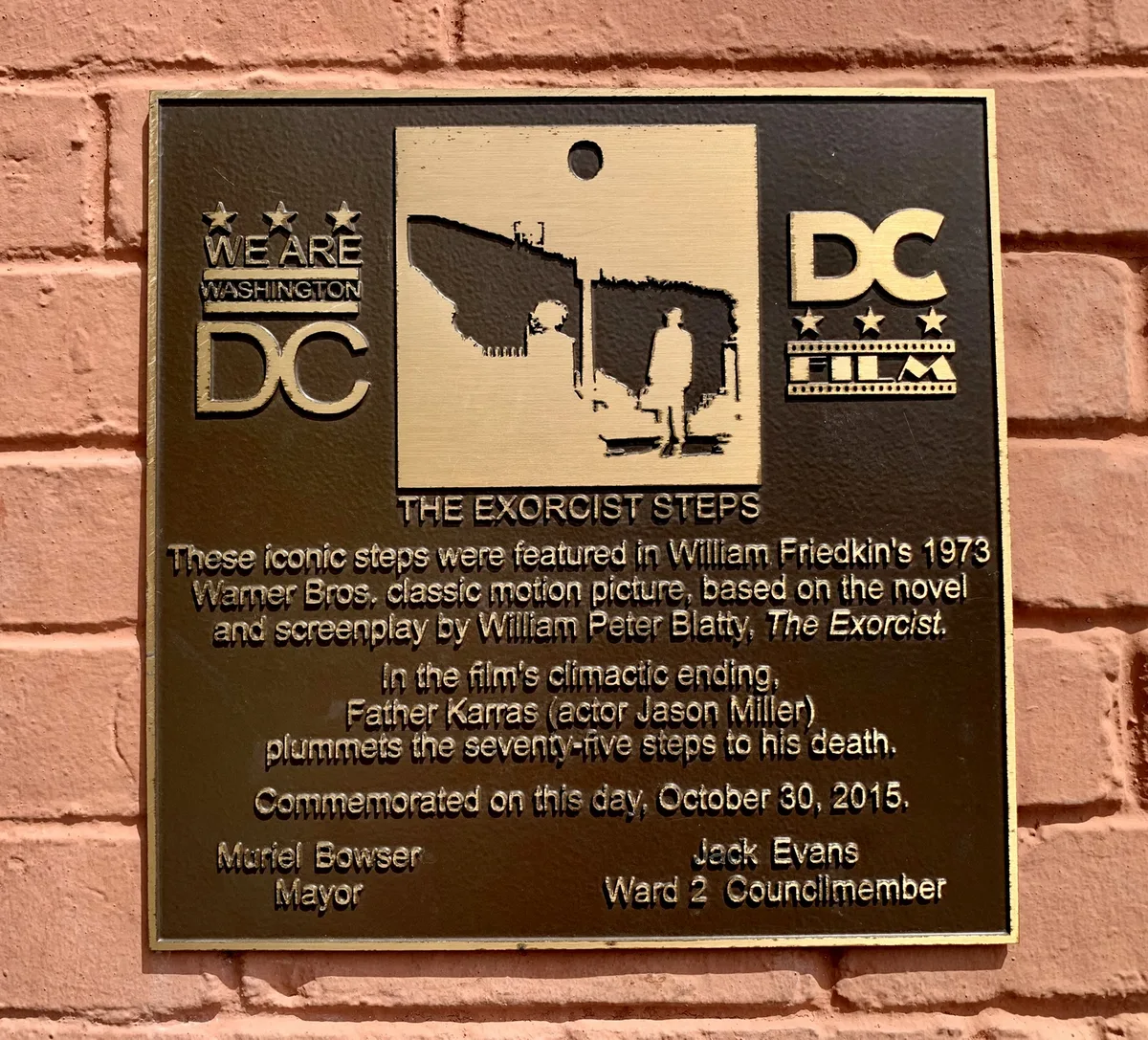 A bronze plaque mounted on a pink brick wall commemorates the Exorcist Steps, with raised DC and “We Are Washington” logos framing a silhouette of the famous staircase. The engraved text is sharply lit, making the plaque’s black-and-gold finish stand out against the warm brick.