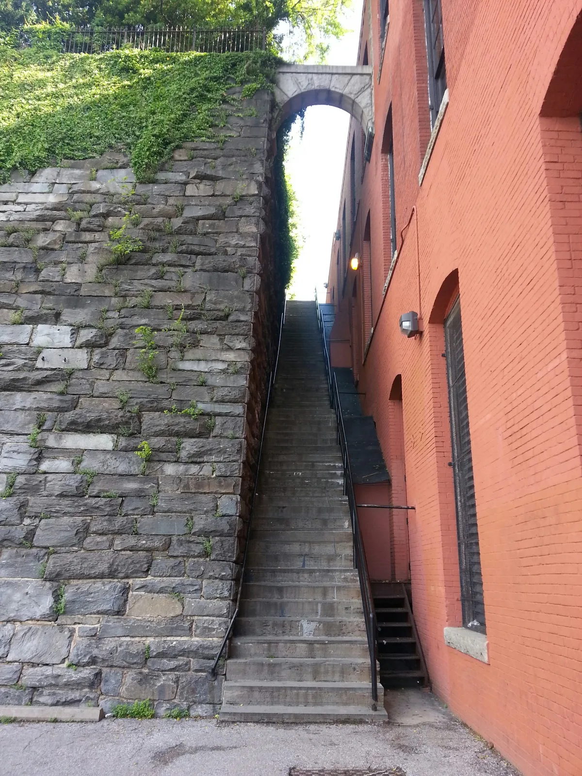 Steep gray stone steps climb between a vine-covered retaining wall and a tall red-brick building, with a stone arch bridging overhead near the top. The narrow passage is lit by soft daylight, emphasizing the dramatic vertical lines and urban film-location feel.