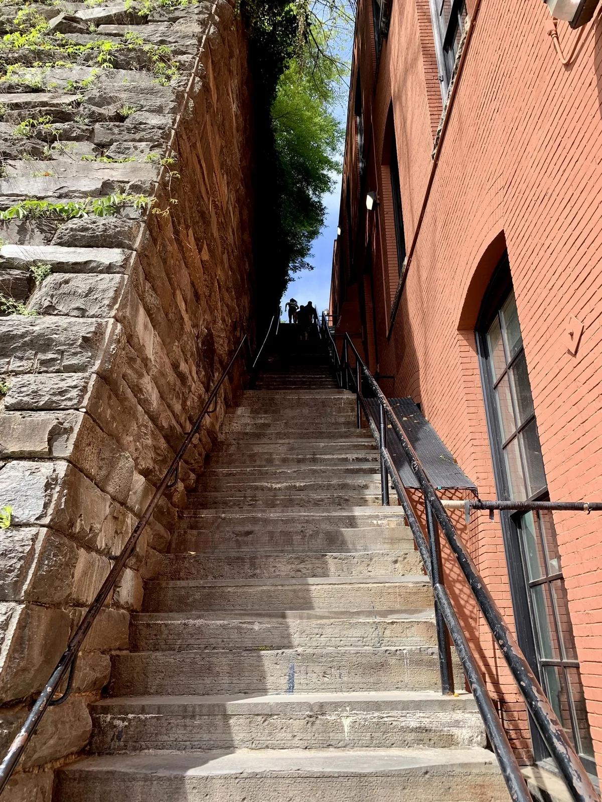 Steep concrete steps climb between a rough stone retaining wall on the left and a tall red-brick building on the right, with black handrails leading the eye upward. Bright daylight and a patch of blue sky frame a few small figures at the top, giving the narrow passage a dramatic, vertical feel.