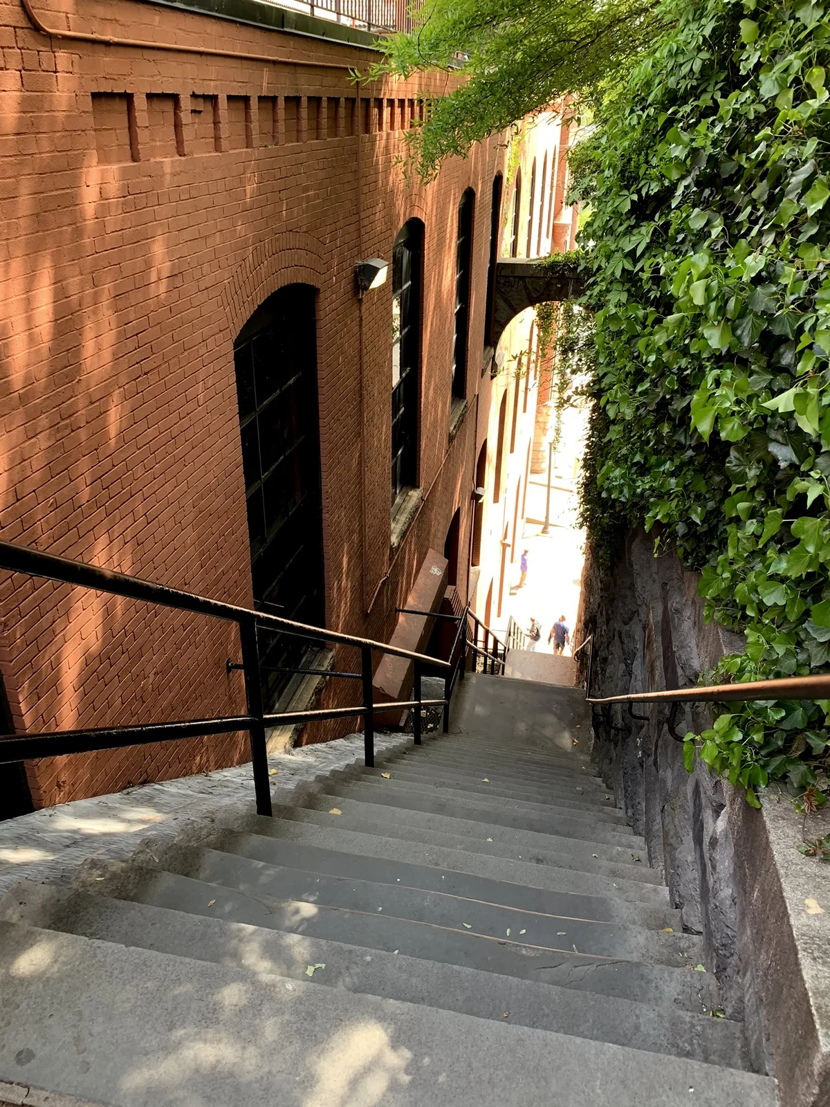 Steep gray stone steps drop between a tall red-brick building with arched windows and a lush ivy-covered wall, with warm golden light brightening the lower landing. Black railings and a few small figures below emphasize the narrow, dramatic urban descent.
