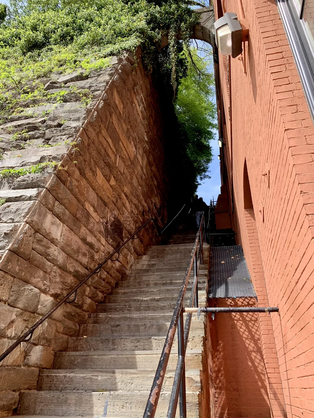 Steep stone steps climb between a rugged retaining wall on the left and a salmon-red brick building on the right, with black handrails leading up toward a bright slice of sky and leafy trees above. Sunlight highlights the textured stone and brick, while the upper landing falls into deep shade.