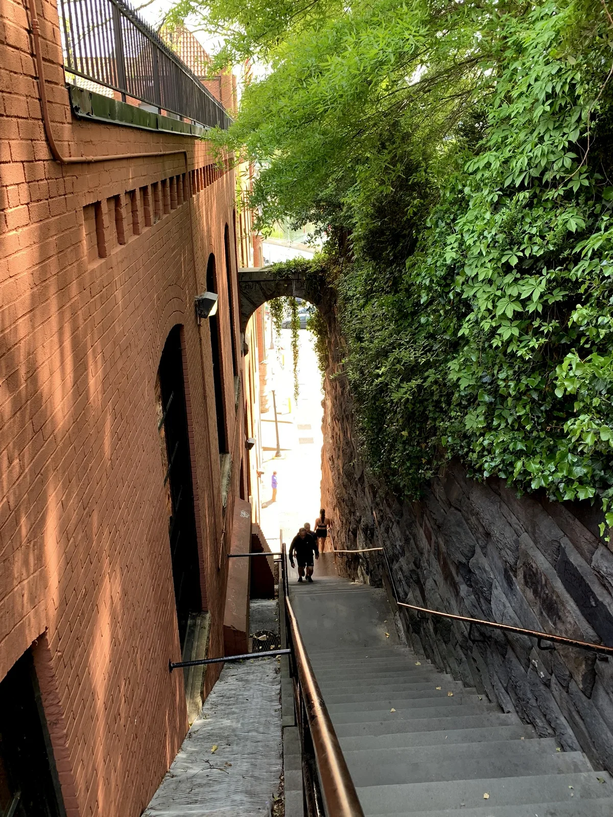 Steep gray stone steps drop between a tall red-brick wall on the left and a lush ivy-covered rock face on the right, with copper handrails guiding the eye down toward a bright opening below. A few people climb the stairs in warm late-day light, emphasizing the narrow, dramatic urban passage.
