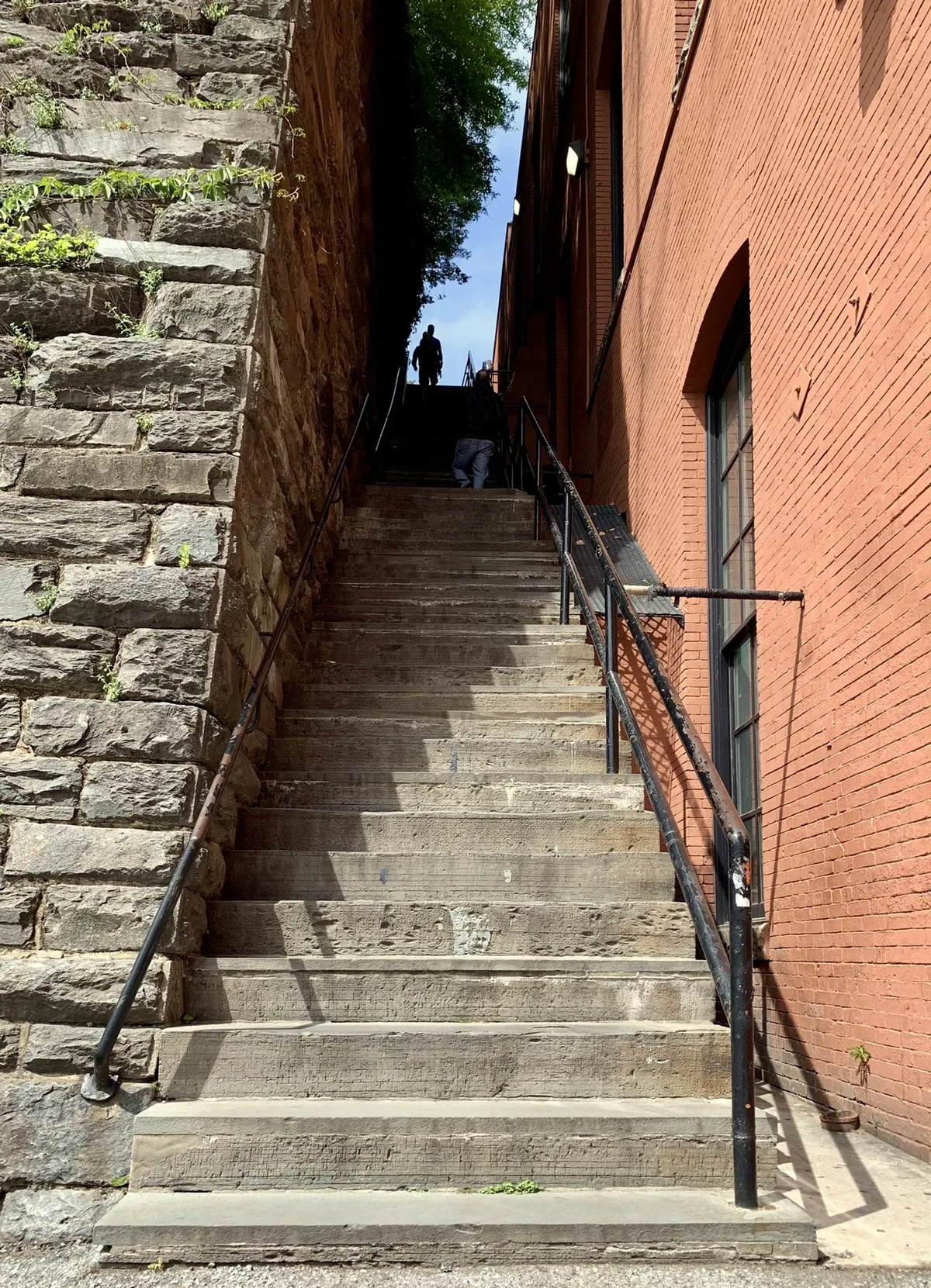 Steep concrete Georgetown Steps rise between a rough stone retaining wall on the left and a tall red brick building on the right, with black iron railings guiding the eye up toward a bright patch of sky. Two small figures stand near the top in deep shade, contrasting with the sunlit lower steps and warm golden-hour light.