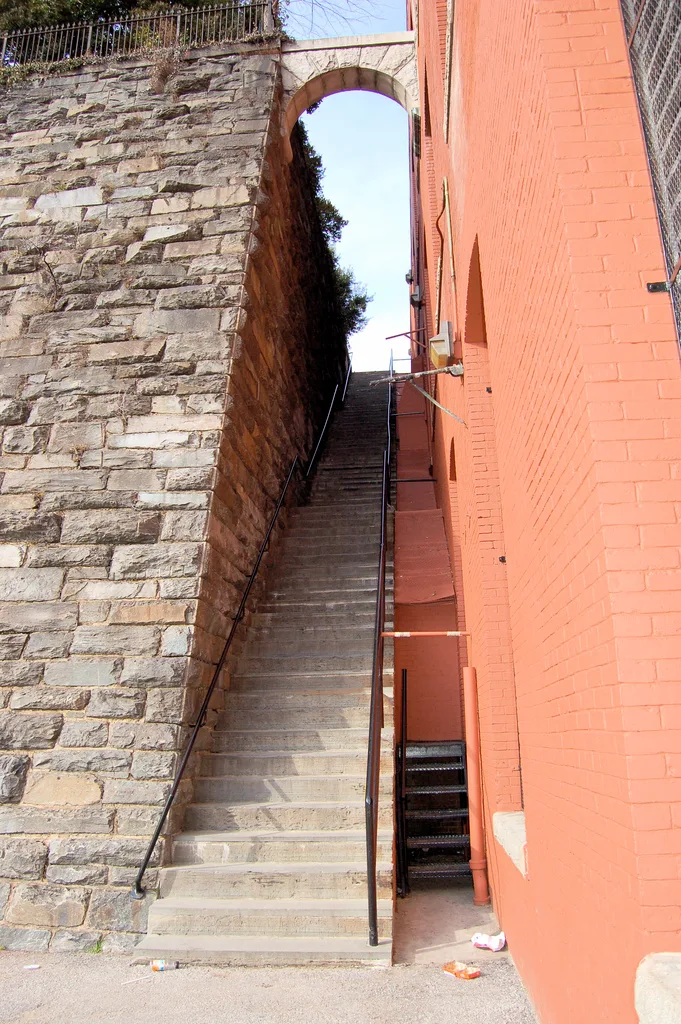Steep gray stone steps rise between a rough stone retaining wall and a salmon-orange brick building, with black railings leading up to an arched opening framed by sky and a bit of greenery above. The tight vertical composition emphasizes the narrow urban passage and the dramatic climb of the Georgetown Steps.