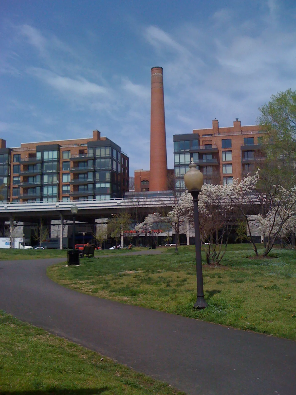 A tall brick smokestack rises between modern brick-and-glass waterfront apartments, with a white bridge railing cutting across the middle of the scene. In the foreground, a curving paved path winds through green grass and flowering trees beneath a soft blue sky.