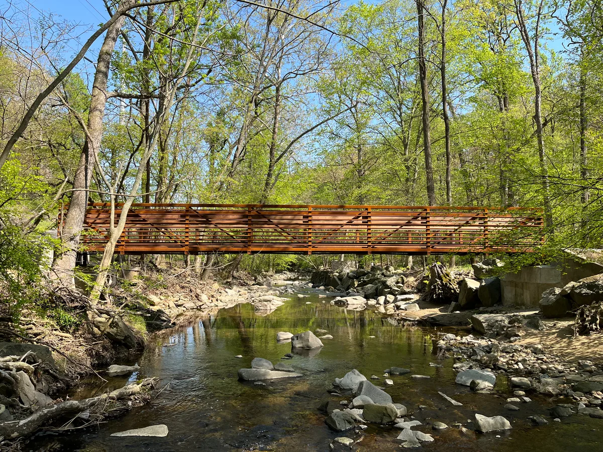 A rust-colored footbridge spans a shallow, rock-strewn creek in a wooded park, with spring-green leaves and tall bare trunks filtering warm sunlight. The calm water reflects the bridge and forest, giving the scene a quiet, tucked-away feel.