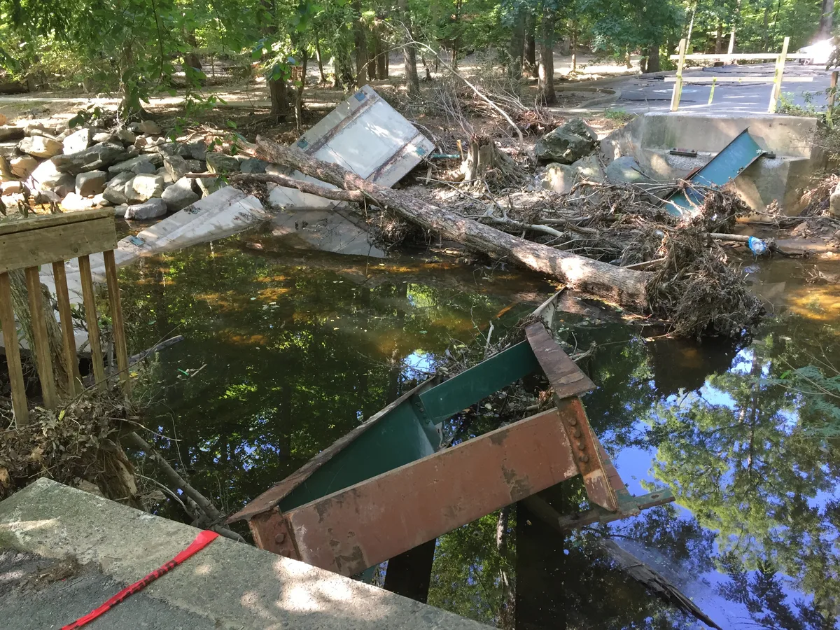 Broken footbridge remains span a shallow, dark creek in a wooded park, with rusted metal beams, concrete rubble, and a fallen log tangled across the water. Sunlight filters through the trees, reflecting blue sky and green canopy on the creek’s surface.