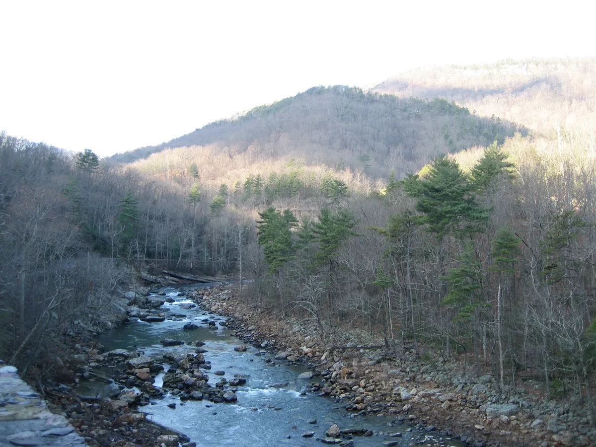 Maury River winds through a rocky gorge, with scattered boulders in the water and leafless trees mixed with evergreens along the banks. Sunlight washes the distant mountain slopes under a pale morning sky.